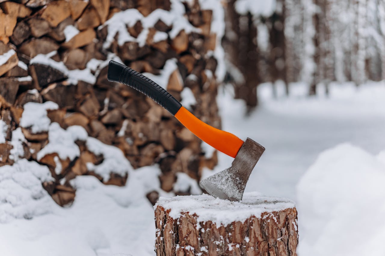 Photo of a wood-chopping axe in a chopping block with a woodpile and snow in the background.